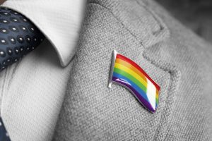 Un homme portant une veste de costume gris assortie d'une cravate bleu marine. Sur la poitrine de sa veste, un pin's en forme de cœur arbore fièrement le drapeau LGBT aux couleurs vives, représentant la diversité et l'inclusivité de la communauté queer.